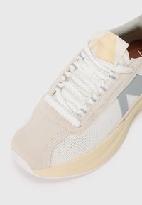 Beige Sneaker aus Wildleder und Mesh-Materialien, mit weißen Schnürsenkeln, grauen Akzenten und einer strukturierten Gummisohle. Sauber sichtbare Nähte.