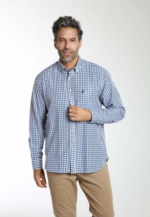 Homme aux cheveux gris portant une chemise à carreaux bleue et blanche à boutons et un pantalon beige, debout devant un fond blanc uni.