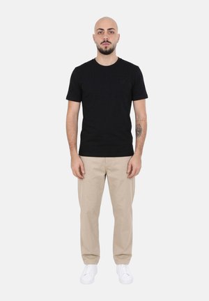 Uomo con barba e tatuaggio che indossa una maglietta nera, pantaloni beige e sneakers bianche, in piedi contro uno sfondo semplice e chiaro.