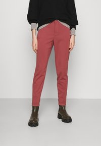 Pantalon rouge effilé en tissu doux, avec une fermeture éclair à l'avant et des poches. Associé à un pull noir et des bottes marron à semelle épaisse.