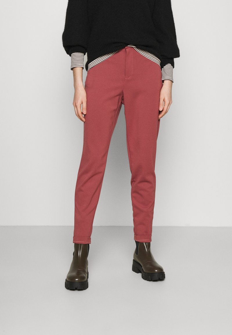 Pantalon rouge effilé en tissu doux, avec une fermeture éclair à l'avant et des poches. Associé à un pull noir et des bottes marron à semelle épaisse.