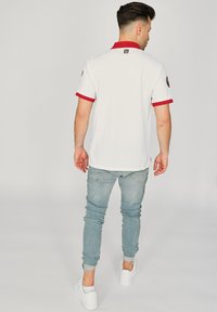 Vit herrpolo med röd krage och ärmslut, med en logotyp på ryggen. Matchas med ljusblå skinny jeans och vita sneakers.