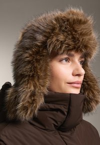 Žena v hnědém zimním kabátu s velkou kapucí olemovanou hustou, načechranou hnědou kožešinou, dívající se doprava.