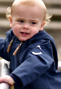 Laivastonsininen takki vetoketjulla, jossa on nahkasisustuksia ja valkoinen logotunnus. Lapsi pitää kiinni kaiteesta, vaaleat kiharat hiukset ja siniset silmät.