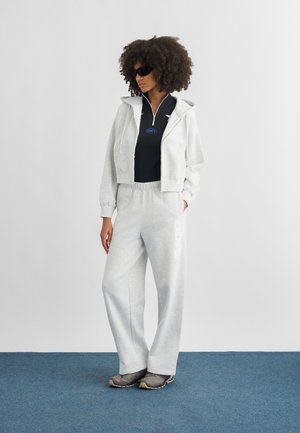 Mujer con cabello rizado que lleva gafas de sol negras, una sudadera negra con cremallera debajo de una sudadera con capucha gris claro, pantalones deportivos grises a juego y zapatillas grises.