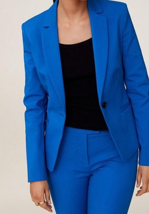 Femme portant une veste de costume bleu vif et un pantalon assorti avec un haut noir, debout devant un fond uni clair.