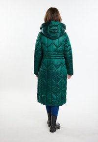 Cappotto verde lungo con imbottitura a diamante, cappuccio e vita aderente. Indossato sopra leggings blu e stivaletti neri alla caviglia.