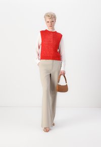Raudona nertinė liemenė ant baltos palaidinės, derinama su smėlio spalvos plačiomis kelnėmis. Laiko mažą rudą handbagą. Elegantiškas, tekstūruotas dizainas.