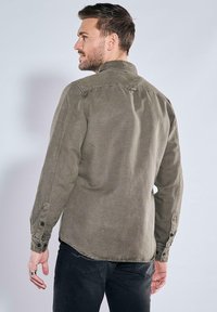 Olivgrünes Hemd mit Knopfleiste, klassischem Kragen, Vordertaschen und sichtbaren Nähten. Hergestellt aus weichem Stoff, mit langen Ärmeln.