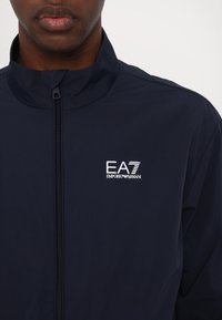 Giacca zip-up blu navy con colletto alto, presenta un logo bianco EA7 Emporio Armani sul petto, realizzata in materiale leggero.
