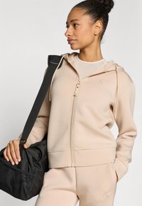 Μπεζ φούτερ με φερμουάρ από απαλό ύφασμα, με διακριτικό λογότυπο, συνδυασμένο με μαύρη τσάντα duffel, που παρουσιάζει ομαλή υφή και λειτουργικό σχεδιασμό.