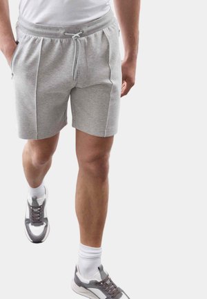 Hombre caminando con pantalones cortos de cordón gris claro, calcetines blancos, zapatillas grises y blancas, y una camiseta blanca sobre un fondo sencillo.