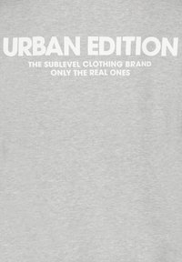 Hellgraues T-Shirt mit fettem weißen Text: "URBAN EDITION" und "DIE SUBLEVEL CLOTHING BRAND NUR FÜR DIE ECHTEN." Einfaches Design, weicher Stoff.