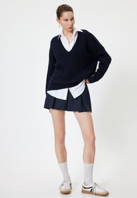 Tummansininen neulepaita V-kaula-aukolla, käytössä valkoisen kauluspaitan ja naviin raidallisen plisessihameen päällä. Asustuksen täydentää valkoiset sukat ja lenkkarit.