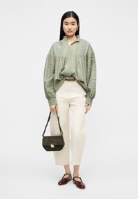 Γυναίκα που φοράει μπλούζα σε χρώμα φασκόμηλου με υφή, κρεμ παντελόνι cropped, καφέ ζώνη, κρατάει μια τσάντα σε σκούρο πράσινο και μπορντό παπούτσια Mary Jane.