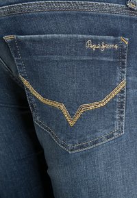 Tmavě modré džíny z denimu s kapsou, která má žluté švy ve tvaru písmene V. Logo "Pepe Jeans" je vyšívané nad kapsou. Hladká textura.