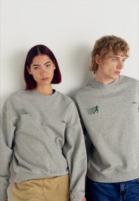 Grijze katoenen sweatshirts met groene accenten en borduurwerk. De ene heeft een lopende figuur; de andere heeft een geometrisch ontwerp op de mouw.