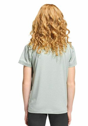 Persona con lunghi capelli biondi e ricci che indossa una maglietta a maniche corte grigio chiaro e pantaloni neri, di spalle su uno sfondo bianco.