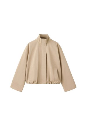 Jeune femme debout portant une veste beige, un crop top blanc, un pantalon blanc, des chaussures marron et un grand sac à bandoulière marron foncé.