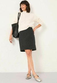 Kreatives Strickpullover in Creme, dunkelgrauer, taillierter Rock und silberne, runde Ballerinas. Die Modell trägt eine schwarze Tote-Bag und steht vor einem hellen Hintergrund.