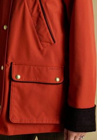 Czerwona wodoodporna kurtka z dużą przednią kieszenią z złotymi zatrzaskami, czarnym zamkiem błyskawicznym i mankietami z kordurowej tkaniny dla dodatkowej tekstury i ciepła.