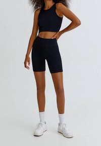 Top corto acanalado negro y pantalones cortos de tiro alto a juego, combinados con zapatillas blancas y calcetines hasta el tobillo, sobre un fondo sencillo.