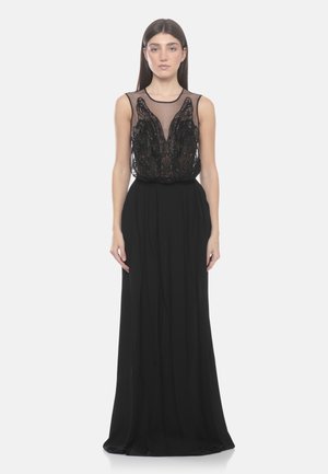 Donna che indossa un abito da sera nero senza maniche con corpetto trasparente ricamato e gonna lunga e fluente, in piedi davanti a uno sfondo bianco.