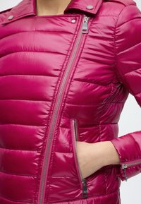 Femme portant une veste matelassée magenta brillante avec une fermeture éclair argentée asymétrique et la main dans la poche latérale zippée.