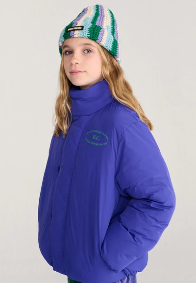 Jeune personne portant une doudoune bleu vif et un bonnet tricoté vert, blanc et violet avec une étiquette "Bobo Choses", debout sur un fond uni.