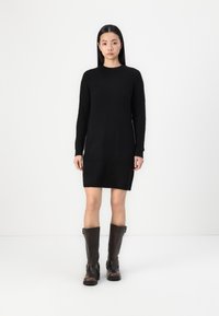 Vestido negro de punto con mangas largas, escote redondo y aberturas laterales; combinado con botas de cuero marrón hasta la rodilla con hebillas.