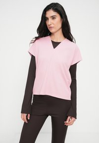 Ροζ κοντομάνικη μπλούζα με ντεκολτέ σε σχήμα V, φορεμένη πάνω από μακρυμάνικο σκούρο καφέ πουκάμισο, από απαλό ύφασμα με άνετη εφαρμογή.