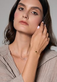 Złota bransoletka z heksagonalnymi akcentami noszona jest na nadgarstku. Modelka ma długie włosy i nosi miękką, teksturowaną beżową bluzkę.