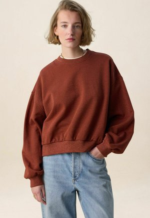 Jonge vrouw met kort blond haar draagt een oversized sweatshirt in roestkleur en lichtblauwe spijkerbroek, staand met één hand in haar broekzak.