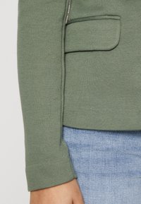Mâneca unui sacou texturat verde, cu un buzunar frontal, asortată cu blugi de culoare bleu deschis. Mâneca are un finisaj curat, bine fasonat.