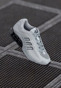 Light gray athletic shoe with mesh upper, three black stripes, and distinctive cushioned sole. Features laces and padded ankle collar.