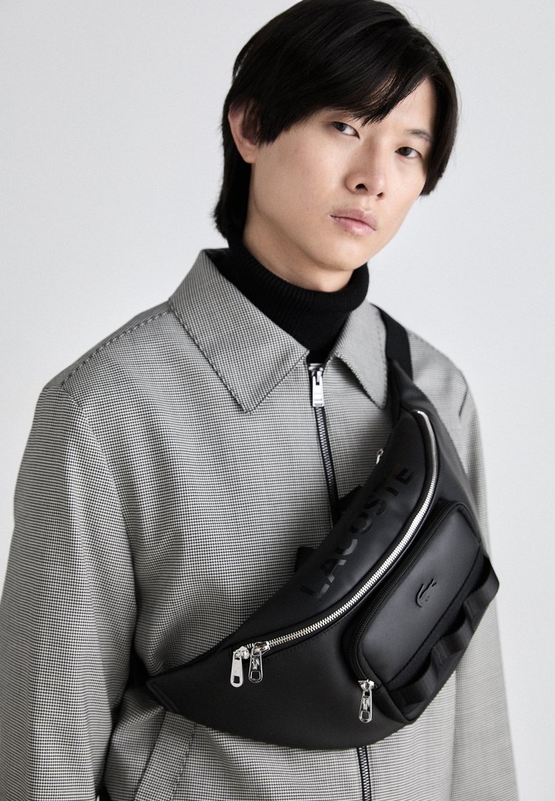 Sac bandoulière en nylon noir avec un design courbé, comportant un compartiment zippé, un logo en relief et une sangle réglable sur un manteau à carreaux.