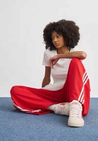 Červené sportovní kalhoty s bílými pruhy, v kombinaci s přiléhavým bílým tričkem a růžovými teniskami, sedící na modrém koberci.