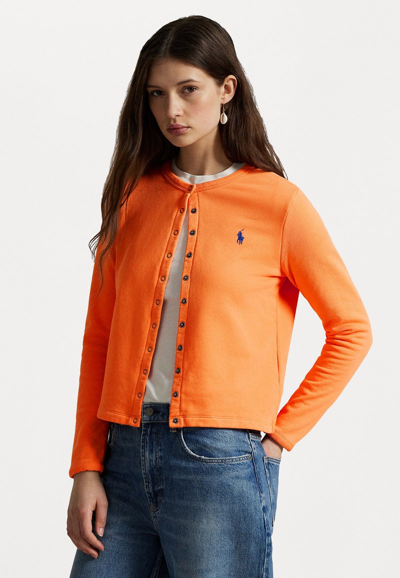 Bright orange cardigan with a round neckline, button closure down the front, and a small blue logo. Worn over a white shirt and denim jeans.