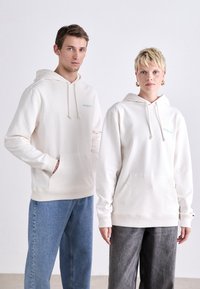 Des sweat-shirts crème clair en tissu doux comportent des poches avant et des cordons de serrage, avec un petit logo vert sur la zone gauche de la poitrine.