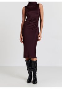 Robe midi sans manches couleur prune foncé avec un détail froncé sur le côté, portée avec des bottes noires brillantes montant jusqu'au genou, sur un modèle debout.