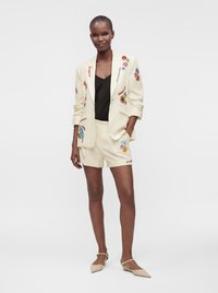 Personne grande portant un ensemble blazer crème et short avec une broderie florale colorée et un haut noir, debout avec une main dans la poche.
