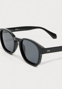 Juodos apvalios saulės akinių su tamsiais lęšiais ir storo blizgaus rėmo, resting ant baltos paviršiaus, su "Revaux" prekės ženklu ant šono.