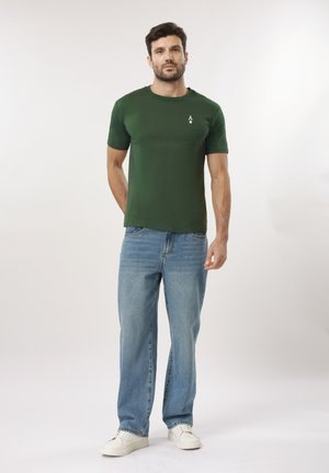 Man standing facing forward wearing a dark green t-shirt with a small white logo, light blue jeans, and white sneakers on a plain background.