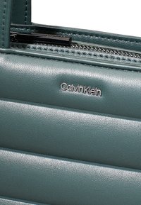 Borsa a mano in pelle teal con texture trapuntata, caratterizzata da un logo argento "Calvin Klein" e una chiusura zip nera nella parte superiore.