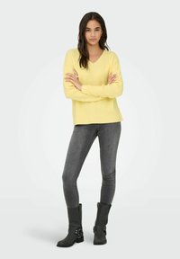 Žuta pletena majica s V-izrezom, dugim rukavima i rebrastom teksturom. Uparena s uskim sivim trapericama i crnim čizmama do članaka s detaljem kopče.