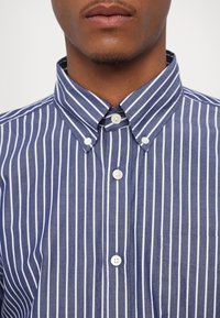Homme portant une chemise bleue à rayures verticales blanches et un col boutonné, visible du cou à la poitrine.