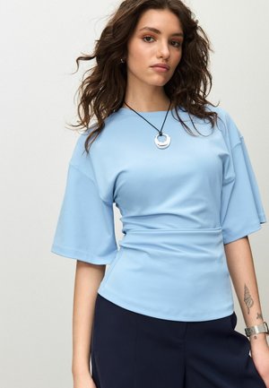 Hellblaues Kurzarmshirt mit tailliertem Schnitt, glatter Textur und rundem Ausschnitt, kombiniert mit dunkler Hose und silberner Anhängerkette.