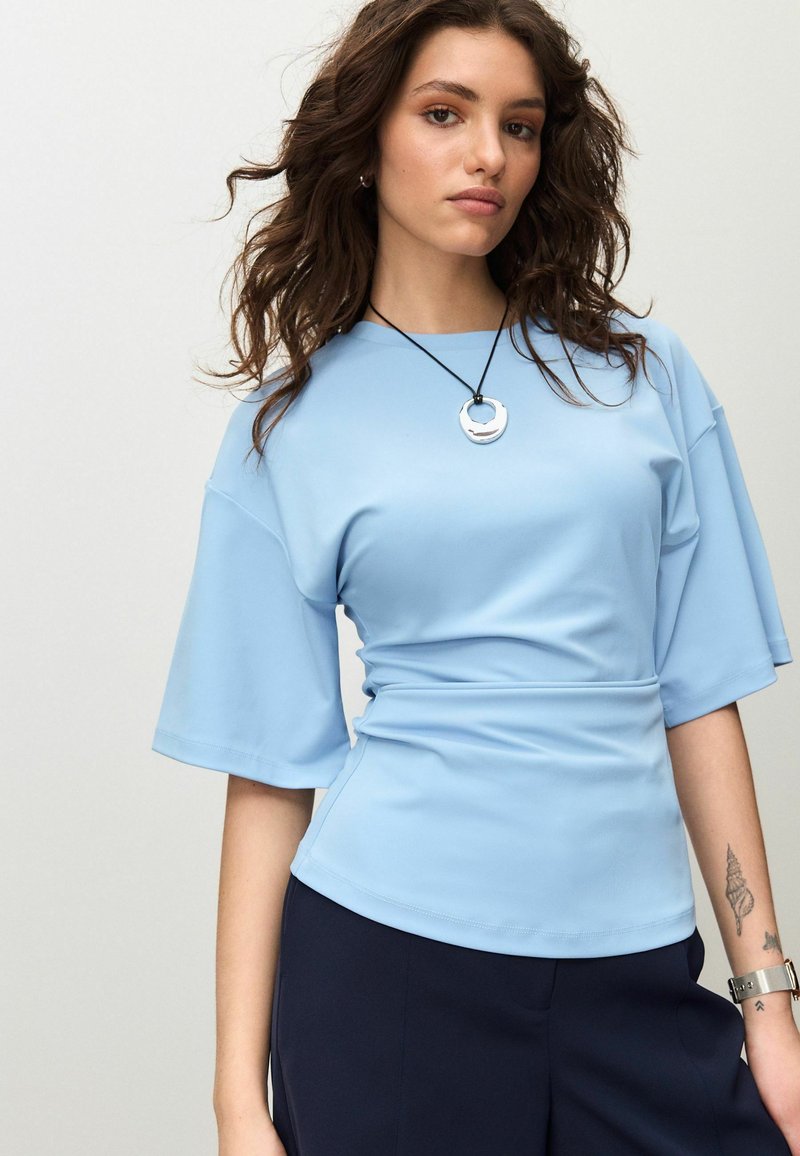 Lyseblå top med korte ærmer og en tætsiddende talje, glat tekstur og rund halsudskæring, kombineret med mørke bukser og en sølvvedhængs halskæde.