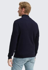 Mann mit blonden Haaren, der einen langärmligen Pulli in Marineblau und helle blaue Jeans trägt, schaut seitlich vor einem schlichten Hintergrund.