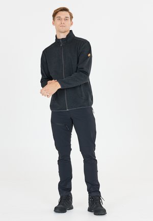 Schwarze Fleecejacke mit Reißverschluss und hohem Kragen, strukturiertem Stoff und seitlichen Taschen, kombiniert mit eng anliegenden dunklen Hosen und schwarzen Stiefeln.